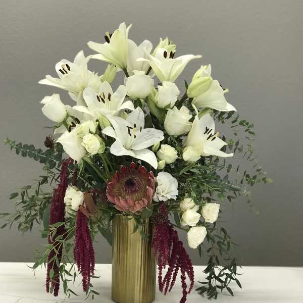 White lilies and roses arranged in a gold vase with burgundy hanging accents.