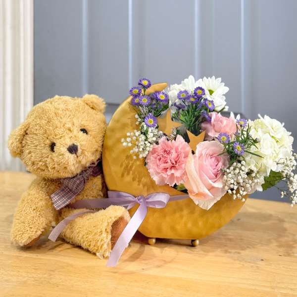 Teddy bear beside a crescent flower arrangement in a gold container