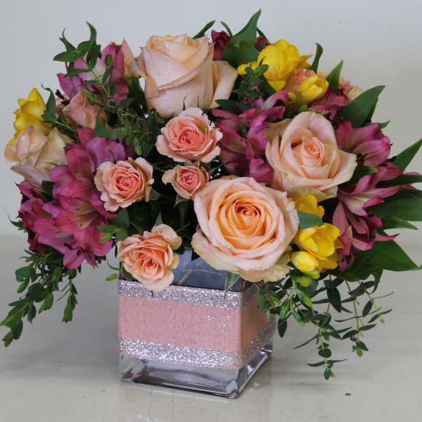 Short arrangement of peach roses, yellow freesia, and pink alstroemeria in a glass cube vase with pink ribbon.