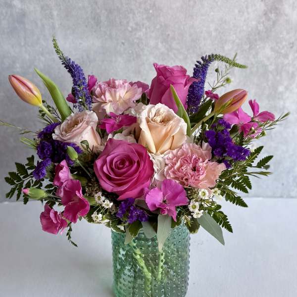 Mixed bouquet of pink roses, carnations, and tulips with purple accents in a textured green glass vase