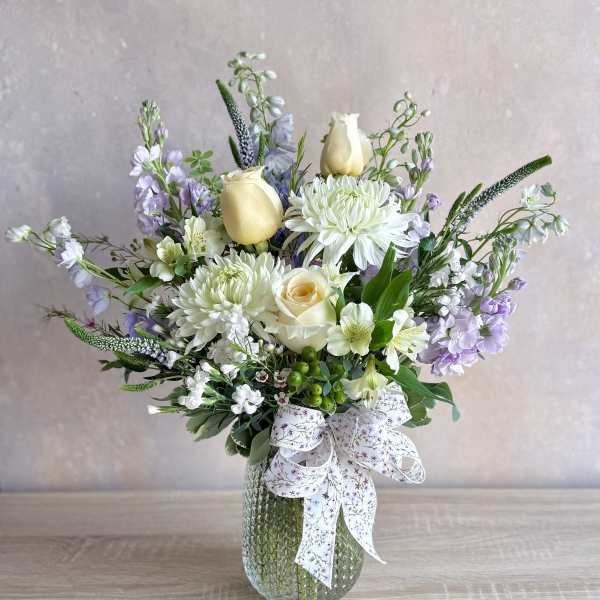 Pastel arrangement of white mums, cream roses, and lavender blooms in a textured glass vase with a ribbon bow.