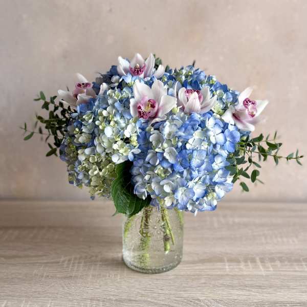 Round arrangement of blue hydrangeas and pink-white orchids in a clear glass jar vase