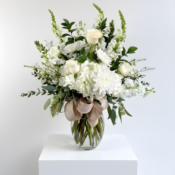 Tall white floral arrangement with roses and mums in a clear glass vase with a beige bow.