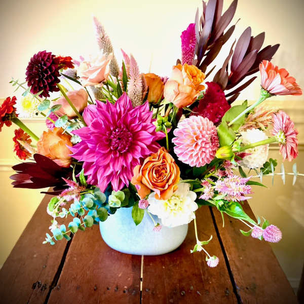 Bright mixed arrangement of pink dahlias, orange roses, and assorted blooms in a white ceramic bowl vase.