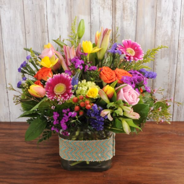 Bright mixed bouquet of roses, gerbera daisies, tulips, and lilies in a square glass vase with burlap band