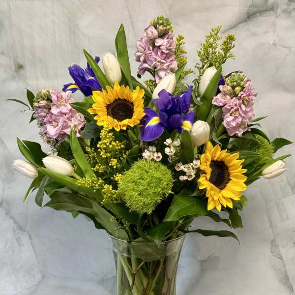 Mixed bouquet with sunflowers, purple irises, white tulips, and pink blooms in a glass vase