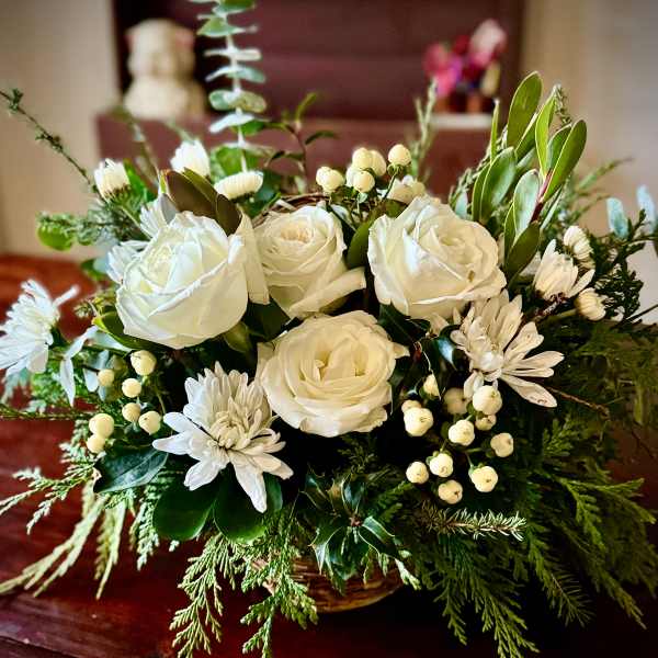 Low arrangement of white roses and mums in a basket with abundant evergreens.