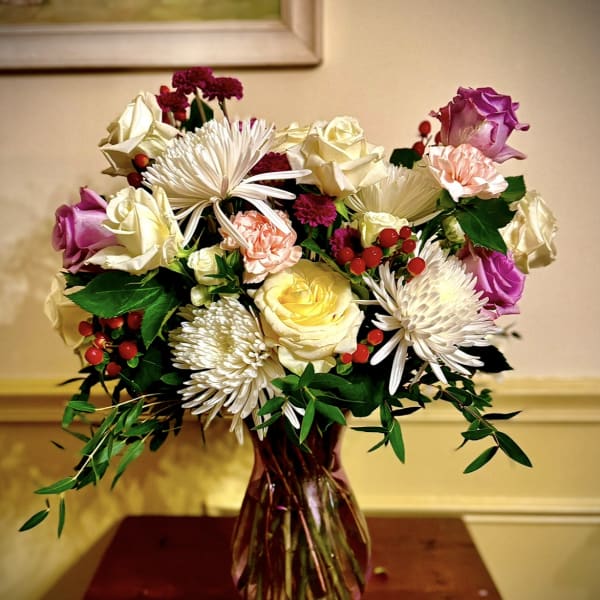 Mixed bouquet of roses, carnations, and white spider mums in a tinted glass vase with red berries