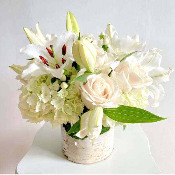 White floral arrangement with roses, lilies, and hydrangeas in a birch-style container