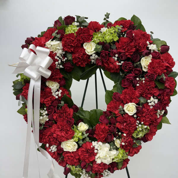 Heart-shaped floral wreath with red carnations and roses, accented by a white ribbon bow