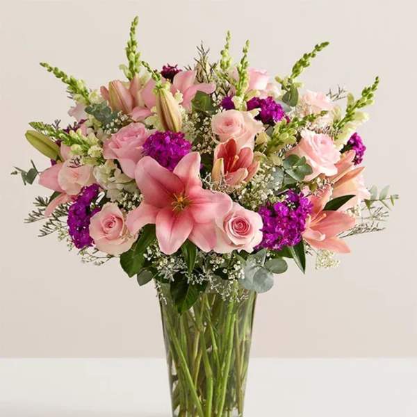 Pink lilies and roses in a clear glass vase with purple accents