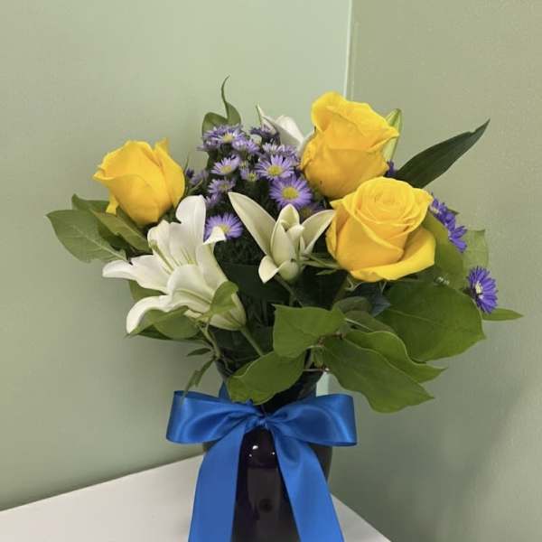 Arrangement of yellow roses, white lilies, and purple asters in a dark vase with a blue ribbon bow