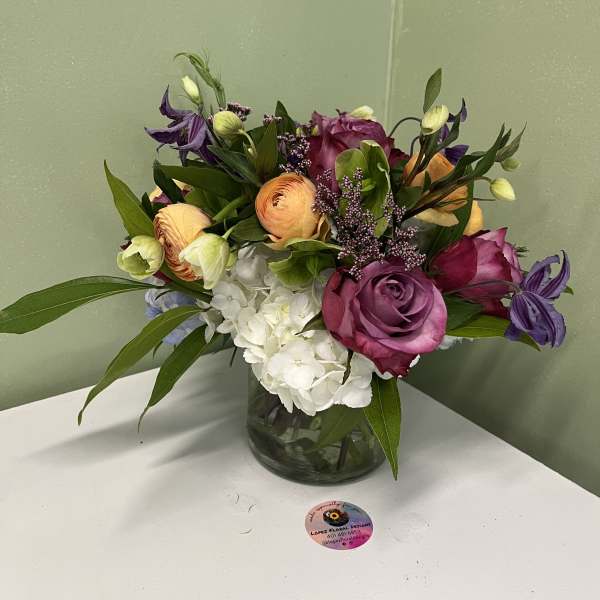 Low arrangement of purple roses, orange ranunculus, and white hydrangeas in a clear glass vase
