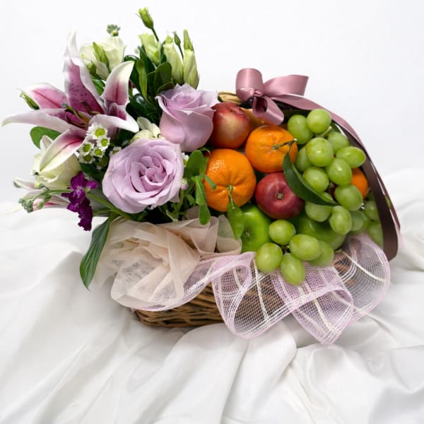 Basket arrangement with lilies, roses, and fruit topped with a pink ribbon