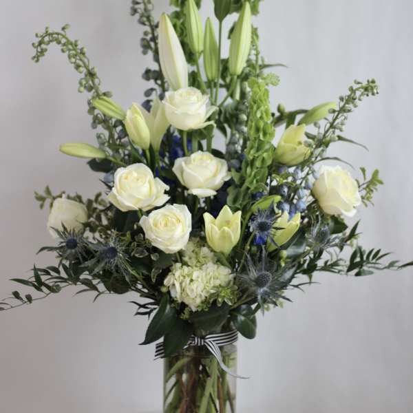 White roses and lilies in a tall glass vase with blue thistle accents