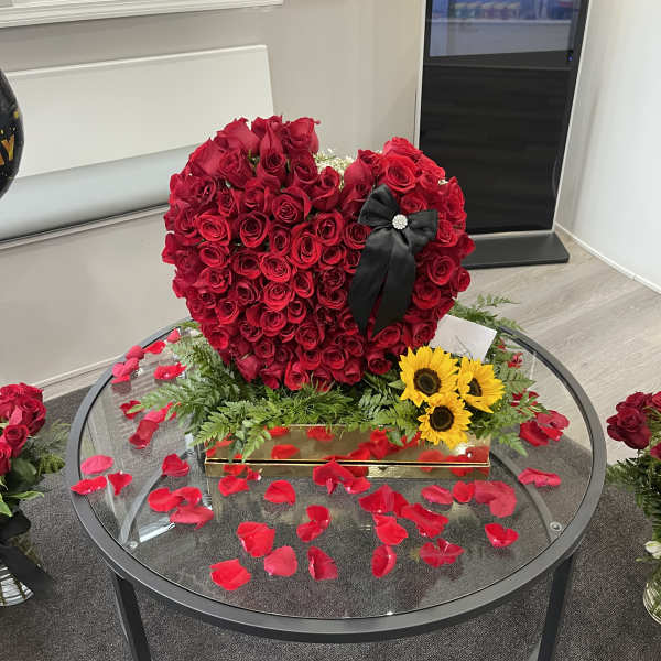 Heart-shaped red rose arrangement with a black ribbon and sunflowers on a glass table