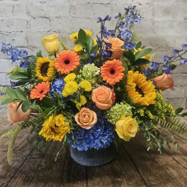 Mixed bouquet of roses, sunflowers, gerbera daisies, and blue hydrangeas in a vase