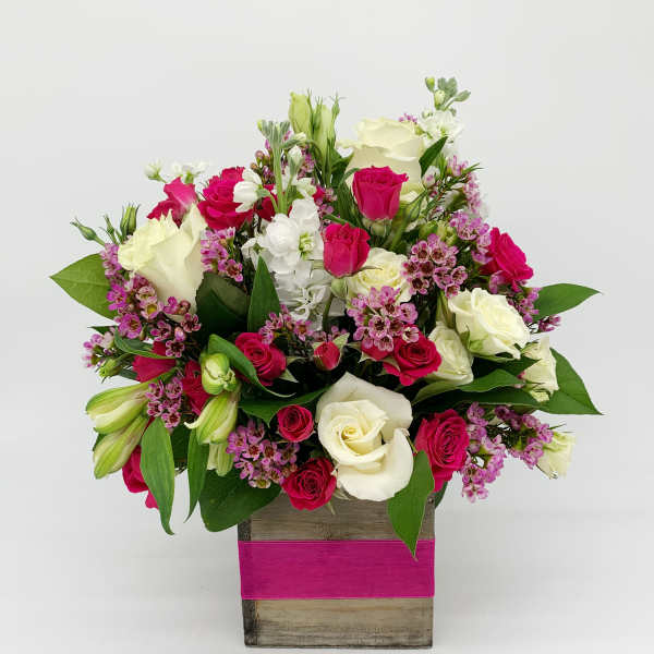 Compact arrangement of white and hot pink roses with small purple flowers in a wood cube vase wrapped with a pink band