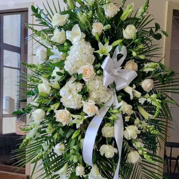 Tall standing spray of white roses, lilies, hydrangeas, and palm fronds with a white bow on an easel.