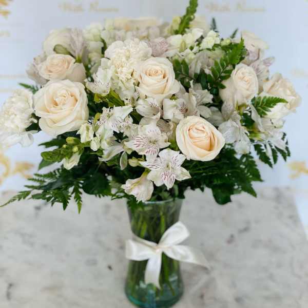 Cream roses and white flowers in a glass vase with a white ribbon