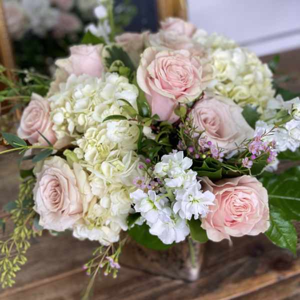 Low arrangement of blush pink roses, white hydrangeas, and white stock in a metallic cube vase on a wooden table.