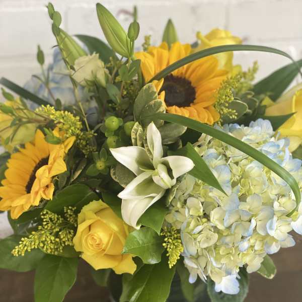 Bouquet of yellow sunflowers, roses, and pale blue hydrangeas in a glass vase