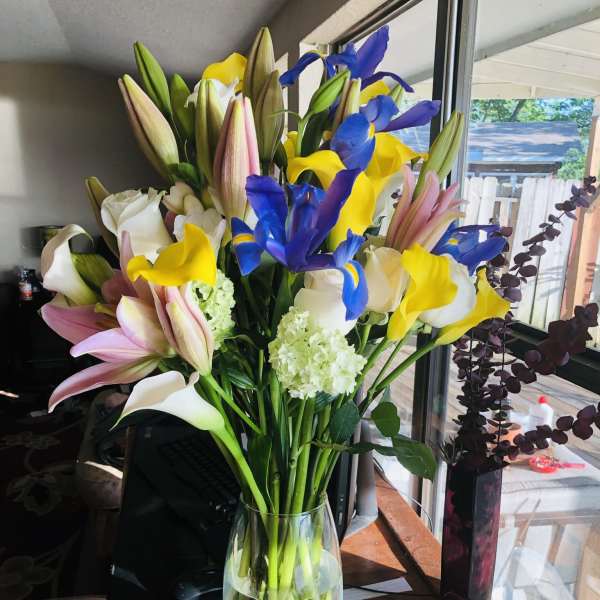 Tall bouquet of yellow calla lilies, blue irises, and pink lilies in a clear glass vase