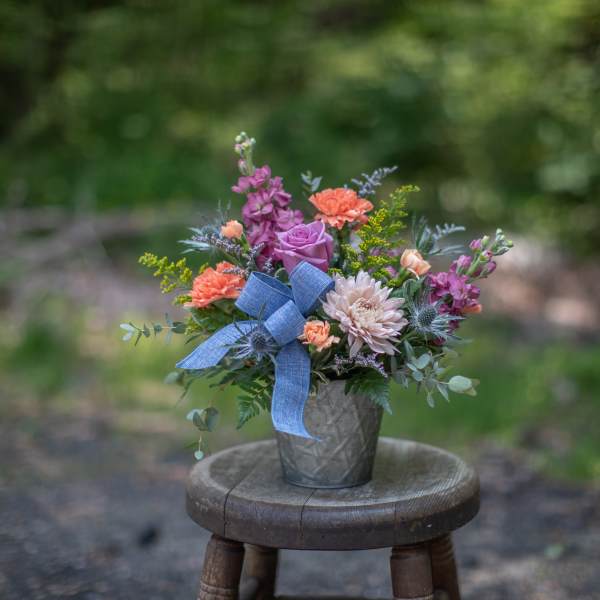 Mixed arrangement of peach carnations, lavender rose, and pink mum in a tin pot with a blue bow.