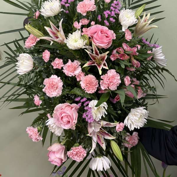 Large standing spray of pink roses, lilies, carnations, white mums, and palm fronds in an oval shape.