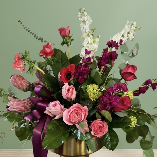 Pink and red mixed flower arrangement in a gold vase with a purple ribbon