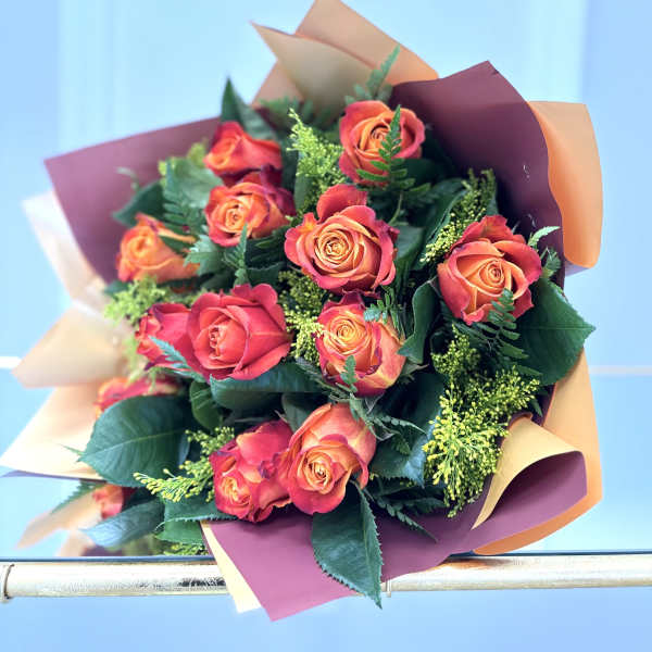 Bouquet of orange and red roses wrapped in peach and burgundy paper against a light blue background
