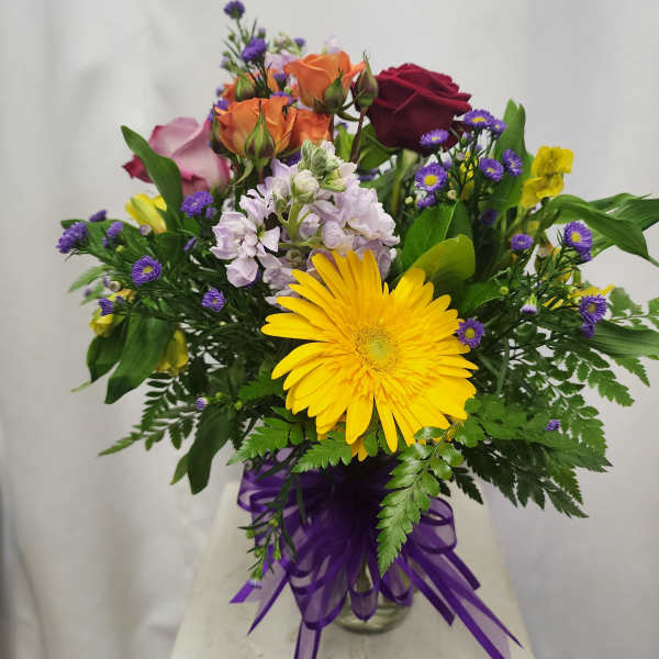 Mixed bouquet with a yellow gerbera daisy, roses, and purple filler flowers in a glass vase