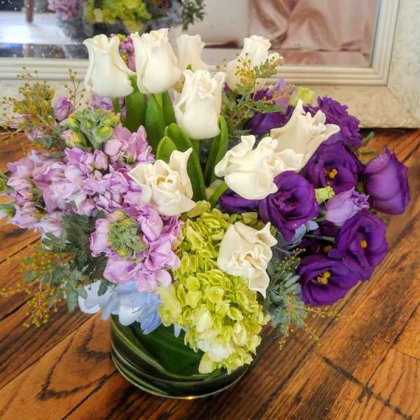 Low round arrangement of white tulips, green hydrangea, and purple lisianthus in a glass vase
