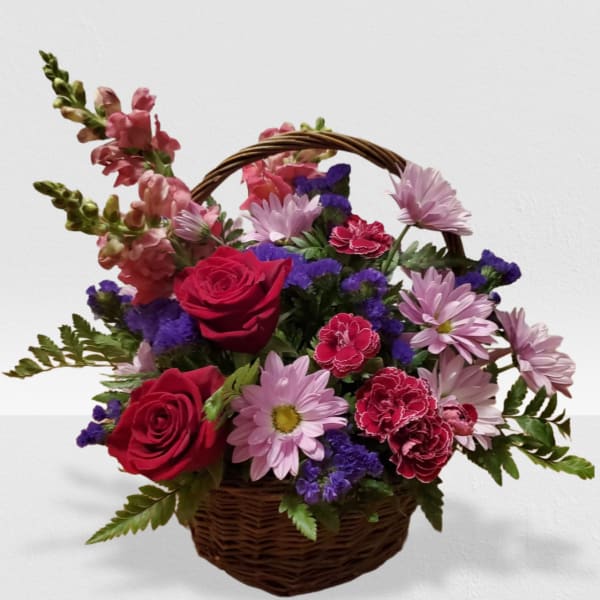 Basket arrangement with red roses, pink daisies, carnations, snapdragons, and purple filler flowers.