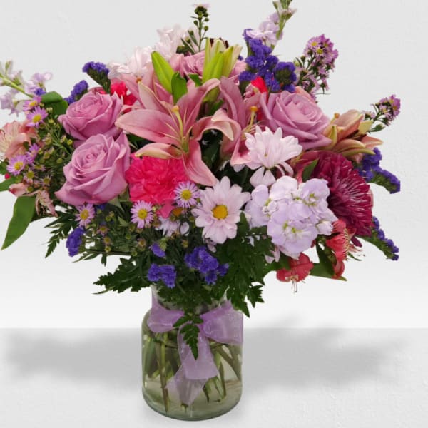 Mixed bouquet of pink lilies, roses, and purple blooms in a clear glass vase with a ribbon bow