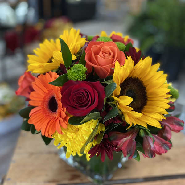 Mixed bouquet of roses, sunflowers, and gerbera daisies in a glass vase