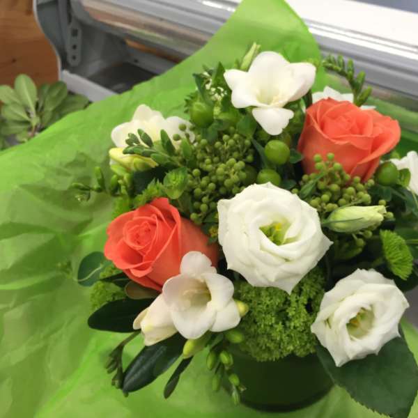 Coral and white roses arranged in a green container with white blooms and greenery.