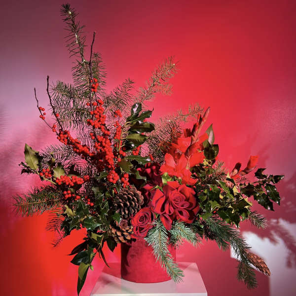 Red roses and evergreen branches arranged in a red vase with pinecones and berries.