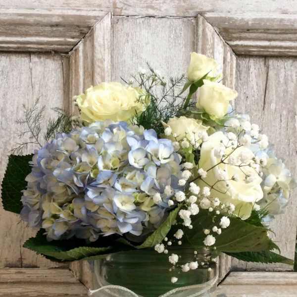 Blue hydrangeas and pale yellow roses in a green glass vase with a ribbon