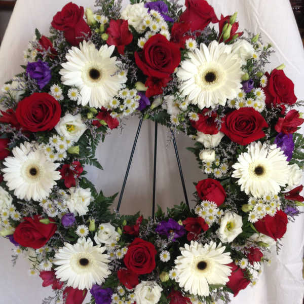 Circular wreath of red roses, white gerberas, and purple blooms on a stand