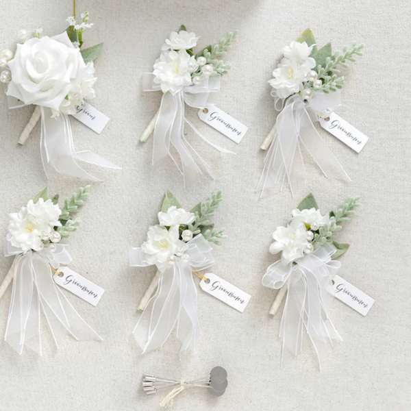 Set of white silk rose boutonnieres with ribbons and groomsmen tags on a light background