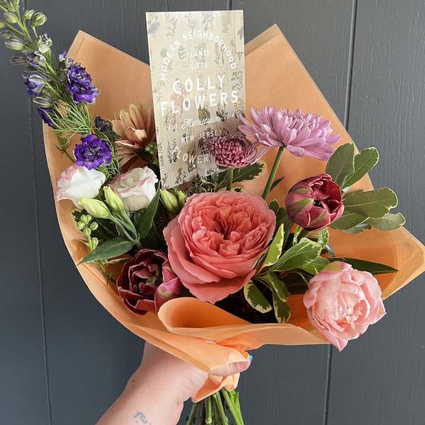 Handheld bouquet of mixed pink, purple, and peach flowers wrapped in orange paper