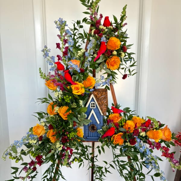 Tall floral easel arrangement with orange roses, blue flowers, and red cardinals