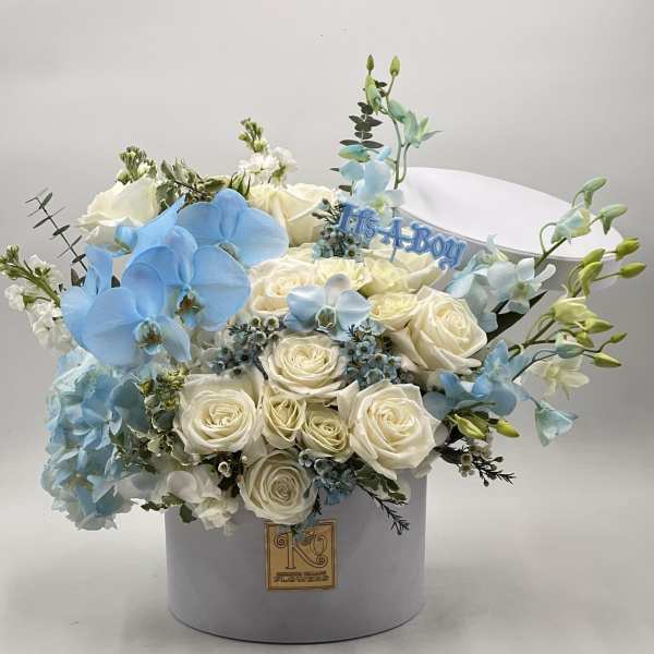 White roses and blue orchids arranged in a round hat box with a "It's a Boy" topper.