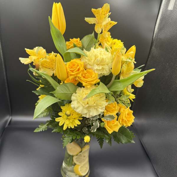 Tall yellow bouquet with tulips, roses, carnations and daisies in a clear vase with lemon slices