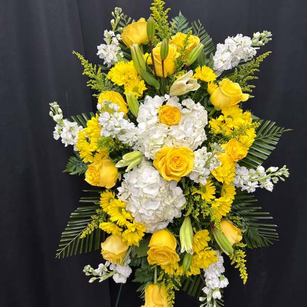 Tall standing spray of yellow roses, white hydrangeas, and yellow daisies on a green easel.