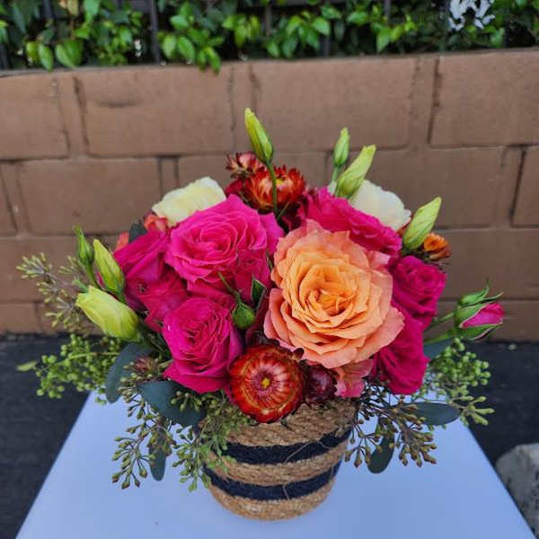 Compact arrangement of hot pink and peach roses in a striped woven basket container