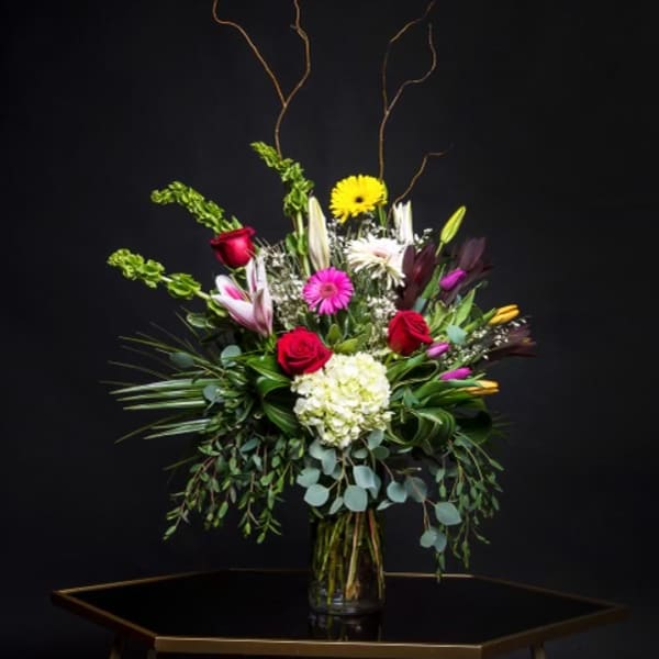 Tall mixed bouquet in a glass vase with roses, lilies, and bright accent flowers