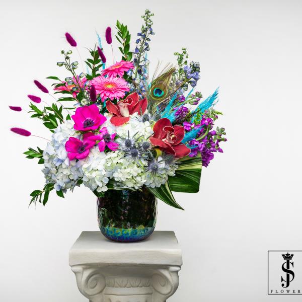 Tall mixed arrangement with white hydrangeas, bright pink blooms, orchids and peacock feather in iridescent vase.
