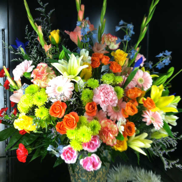 Tall, colorful mixed flower arrangement with roses, lilies, gerbera daisies and carnations in a patterned vase.